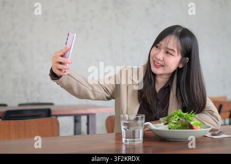 A horizontal composition featuring a young Asian woman holding up a pink smartphone to take a selfie while smiling warmly. She is leaning on a wooden Stock Photo