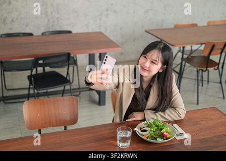 A horizontal, high-angle composition capturing a young Asian woman as she holds out a pink smartphone to take a selfie. She is dressed in a profession Stock Photo