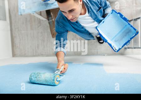 Millennial man paints wall with roller and holds tray in his hand Stock Photo