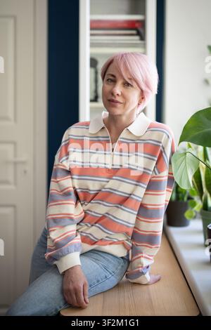 Relaxed positive stylish mature woman posing at camera while sitting on table beside window at home. Stock Photo