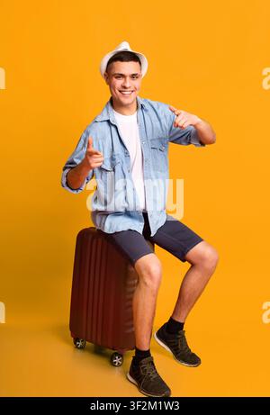Traveler young man indicating at camera, sitting on luggage Stock Photo