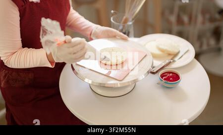 Hands piping frosting onto round cake on turntable with jam bowl and plates on white table in baking workspace Stock Photo