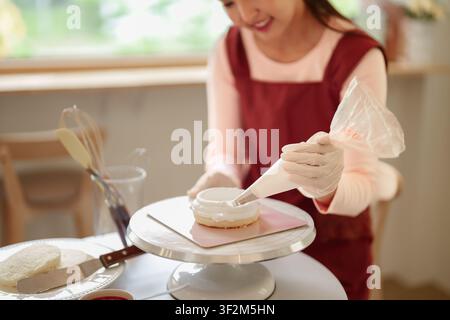 Woman wearing disposable glove decorating homemade cake with cream icing on stand in bright kitchen workspace Stock Photo