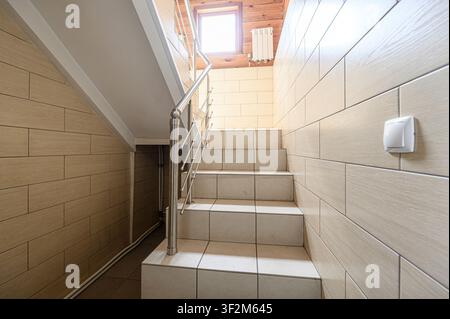 staircase ascends with tiled steps and a stainless steel railing. Walls are covered in light-colored, wood-grain tiles. A small window is visible at t Stock Photo