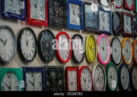 A wall displays rows of analog clocks, varying in shape and color, including circular and square designs with plastic and metal frames. Clock faces ar Stock Photo