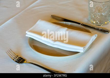 A neatly folded cloth napkin rests on a ceramic plate beside a knife, fork and clear drinking glass Stock Photo