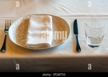 Restaurant place setting with ceramic plate folded napkin drinking glass and cutlery prepared for guest dining experience. Stock Photo