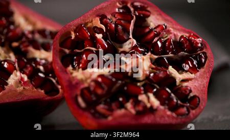 A sliced juicy pomegranate displays tightly packed glossy crimson seeds on a dark background. Deep shadows and intentional low key lighting emphasize Stock Photo