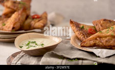 Deep fried triangular pastries sit on parchment paper alongside a small bowl of creamy white sauce decorated with fresh parsley Bright natural light e Stock Photo