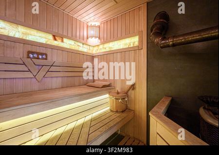 warm, inviting sauna interior with wooden benches, paneling, a bucket, and unique lighting. A rustic pipe detail adds character Stock Photo