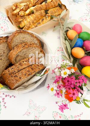 Traditional Easter breakfast with sliced whole grain bread, painted boiled eggs, olive branch and spring flowers on floral tablecloth. Overhead view. Stock Photo