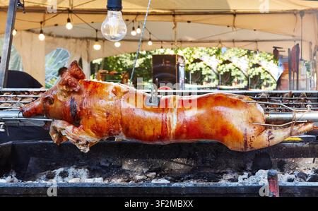 Whole pig is being roasted on a spit over a charcoal grill, located under a tent with decorative lights, creating a lively outdoor cooking atmosphere Stock Photo
