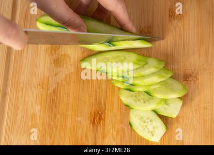 Slicing cucumbers with a large knife on wood cooking board close-up. Making a fresh vegetable salad, a woman cooks at home Stock Photo