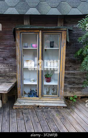 Old distressed wooden cabinet with glass doors stands on rustic wooden deck against weathered wall with shingles. Inside shelves hold colorful bowls c Stock Photo