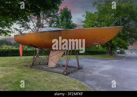 Old orange sailboat hull rests on metal wheeled stand in outdoor park setting. Weathered rusty surface shows signs of age with graffiti markings. Dram Stock Photo