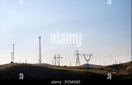 Power lines and towers stretch across a dry, grassy hillside under a clear sky. Several utility poles carry high-voltage cables across the terrain Stock Photo