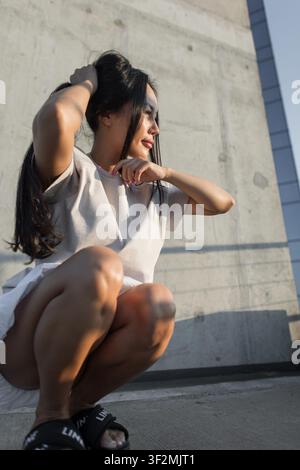 young Asian woman in a casual beige t-shirt squats against a textured concrete wall, smiling and touching her long dark hair in warm afternoon sunligh Stock Photo