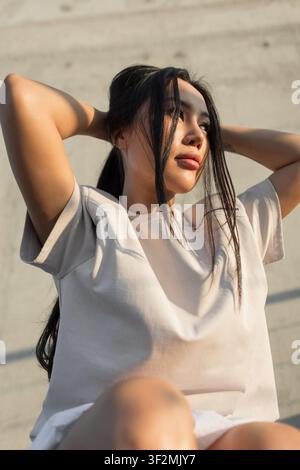 confident young Asian woman in a casual beige t-shirt holds her long dark hair back, looking away into the warm afternoon sunlight against a concrete Stock Photo