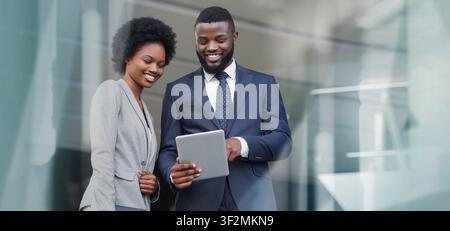 Smiling Black Man And Woman Looking At Tablet In Modern Office, Business Concept Stock Photo
