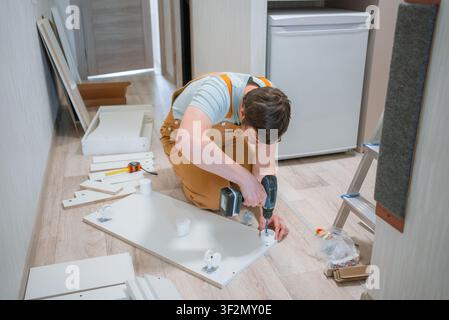 Man kneeling on the floor, carefully assembling a chest of drawers using a battery-powered drill, surrounded by unassembled furniture pieces and tools Stock Photo