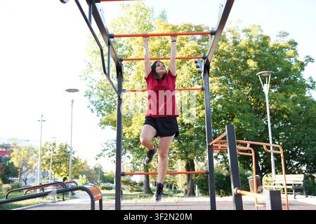 Young woman hanging from monkey bars during outdoor calisthenics workout Stock Photo