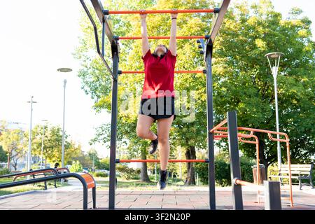 Young woman hanging from monkey bars during outdoor calisthenics workout Stock Photo