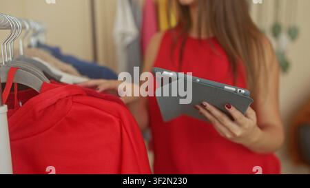 Woman holds tablet and touches clothes with manicured hands in a walkin closet studio wearing red top; thoughtful outfit choice. Stock Photo