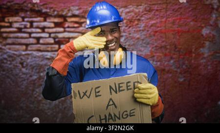 Man in blue hardhat and yellow ear defenders holding a cardboard sign reading 'we need a change' while touching his face with a gloved hand on a stree Stock Photo