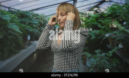 Woman with raised hands and open mouth looking up in a plant filled building aisle, wearing houndstooth top and jeans; wonder discovery nature. Stock Photo