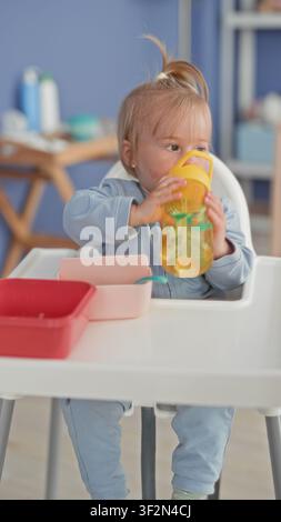 Toddler girl drinking from yellow sippy cup, holds cup to mouth in kitchen highchair with bowls and spoon on tray; calm domestic morning. Stock Photo