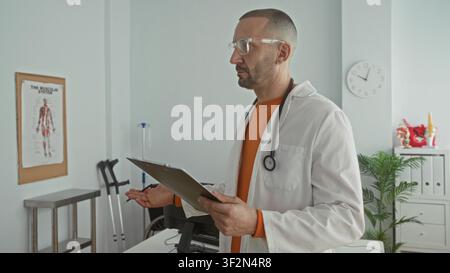 Man doctor with stethoscope and clipboard reads and writes notes in clinic building beside crutches, anatomy poster and wall clock; focused care. Stock Photo