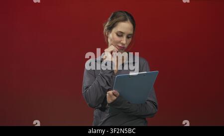 Woman holding a blue clipboard touches her chin in studio with red backdrop and casual sports top; thoughtful reflection. Stock Photo