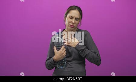 Woman clutching chest while holding a large water bottle in studio, pained expression suggesting discomfort; distress anxiety concern. Stock Photo