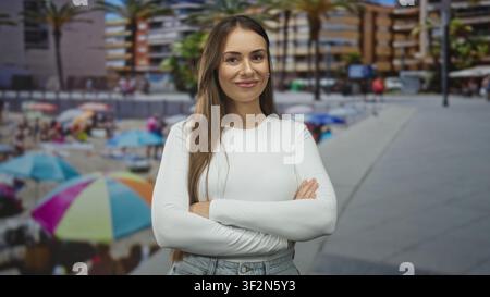 Woman with arms crossed and slight smile wearing a white long sleeve top on a street promenade by beach umbrellas and palm trees; confidence empowerme Stock Photo