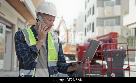 Man builder in hardhat and hi vis vest talks on phone while holding laptop on an urban street with excavator behind him; focus duty. Stock Photo