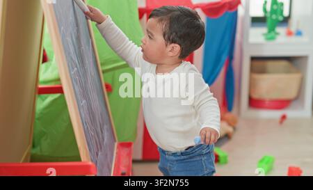 Boy drawing on chalkboard holding chalk and scribbling at an easel in a colorful playroom inside a building; focus learning curiosity. Stock Photo