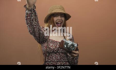 Young woman holding vintage camera with leather case, waving right hand and smiling while wearing straw hat and floral dress in studio; joyful spontan Stock Photo