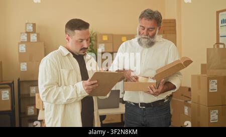 Two men inspect and hold a parcel box while one reads a clipboard in a small business building; teamwork collaboration efficiency. Stock Photo