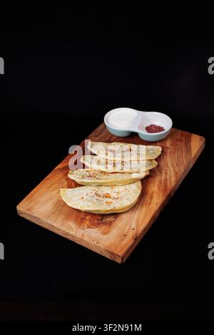 Studio shot of four folded quesadilla tortillas sprinkled with spices, served with dual dipping sauces in a white bowl on a rustic wooden board agains Stock Photo