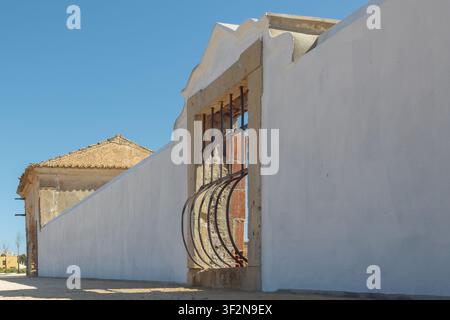 A wall stands beside an old building with a roof. Openings in the wall show a view of the sky. The scene is in a rural area on a clear day. Stock Photo