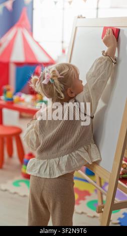 Girl erasing whiteboard on a wooden easel in a playroom near a red tent and colorful playmat, wiping with a red eraser and hand motion; focused learni Stock Photo