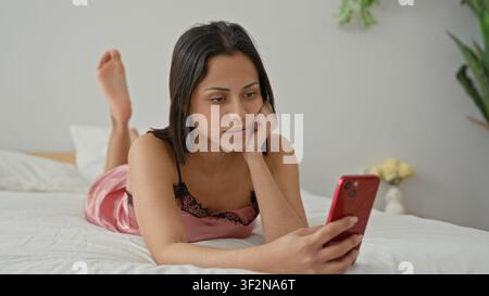 Young woman on bed in pink nightdress holding smartphone with hand on cheek and legs raised, building; quiet contemplation. Stock Photo