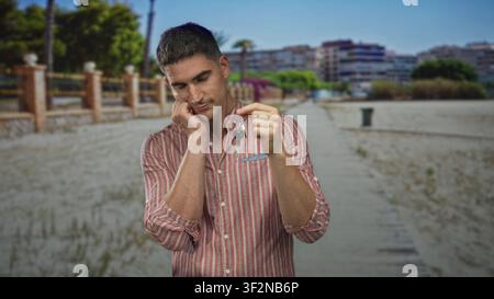Man in striped shirt holding keys on keyring with hand on cheek on street looking down, pensive expression; lost home sadness. Stock Photo