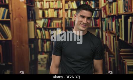 Young man smiling and slightly leaning by library shelves filled with books in a building; calm curiosity. Stock Photo