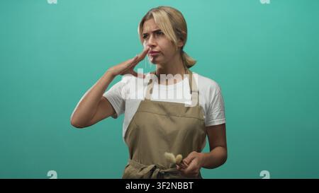 Woman makeup artist young blonde wearing apron pinches nose while holding brush in studio with teal backdrop; disgust reaction. Stock Photo