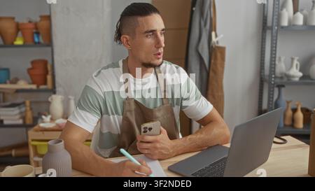 Man in apron holding phone and writing with hand on notepad beside laptop and ceramic vases on shelves in pottery studio; focused creativity. Stock Photo