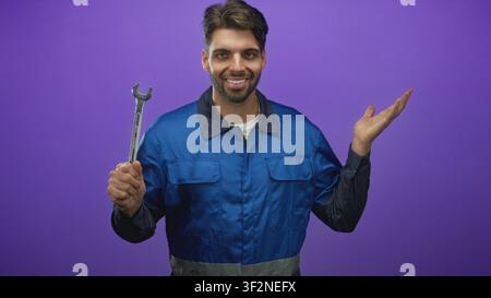 Man mechanic in blue work overalls holds wrench and gestures open palm in a studio setting with purple backdrop; approachable confidence. Stock Photo