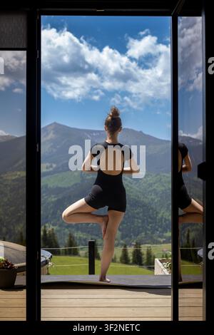 Woman Practicing Yoga with Mountain View. Back view of a woman in black activewear holding a yoga tree pose on a wooden terrace. Stock Photo