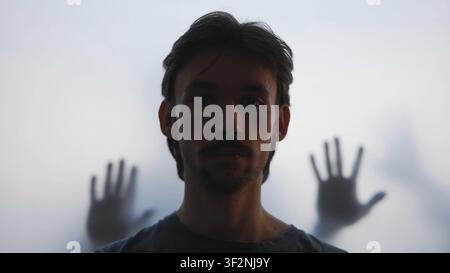Young man in frontal silhouette with blurred hands pressing against frosted panel, creating dramatic indoor contrast, translucent light, tension Stock Photo