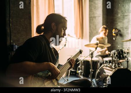 Bass guitarist practicing during recording studio rehearsal, focused musician playing electric bass while drummer performs in background, independent music production. Stock Photo